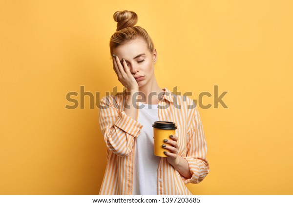 Tired sleepy woman\
holds a cup of coffee, has sad expression, closes eyes, cannot wake\
up in the morning and go to work. difficult, hard monday. isolated\
yellow background