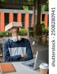 A tired, sleepy young Asian male college student rests with a book covering his face while working on homework or preparing for an exam at an outdoor table.