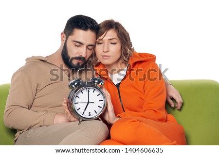 Tired sleepy young couple in home clothes sitting on sofa with closed eyes, with big alarm clock on knees