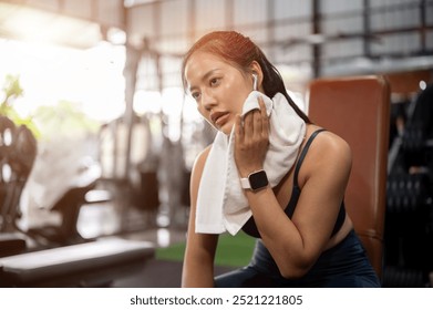 A tired, sporty Asian woman in athletic wear sits on a bench press to rest after her workout in the gym, wiping sweat from her face with a towel. workout, fitness training, exercise - Powered by Shutterstock