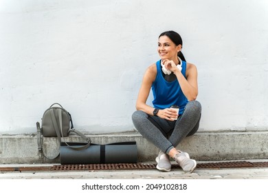 Tired Sporty Woman Resting After Morning Yoga While Holding A Cup Of Coffee And Sitting At The Border Near The Grey Wall Outdoors. Stock Photo 