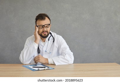 Tired Stressed Doctor Having A Headache On A Bad Day At Work. Studio Portrait Exhausted Young Man In White Lab Coat Touching Head And Rubbing And Massaging Temples Trying To Get Rid Of Terrible Pain