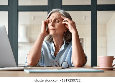 Tired Stressed Old Mature Business Woman Suffering From Headache At Work. Upset Sick Senior Middle Aged Lady Massaging Head Feeling Migraine From Overwork Or Menopause Using Computer At Home Office.