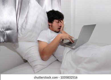 Tired stressed young Asian man feeling sleepy while using laptop on the bed in bedroom.  Hard work concept.