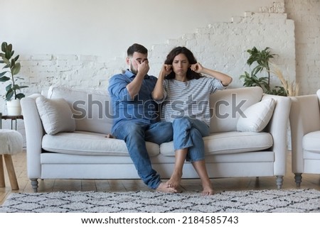 Tired stressed young couple arguing at home, sitting on sofa together. Stubborn wife plugging ears for stop listening to exhausted annoyed husband speaking. Marriage crisis, conflict concept