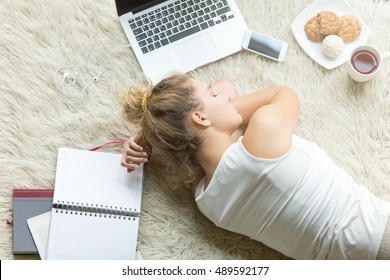 Tired student girl fell asleep after studying hard, sleeping beside laptop, open copybook, cookies and cup of tea on white floor carpet in living room. Education, exams, school concept. Top view