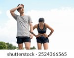 A tired, sweaty Asian couple in sportswear wipes sweat from their faces, taking a break after a long run together in a park in the morning. outdoor activity, sport, healthy lifestyles