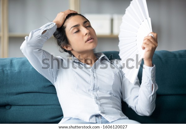 Tired sweaty young indian woman holding using\
hand fan sit on couch in uncomfortable hot summer weather suffer\
without air conditioner cooling feel hot at home apartment,\
overheating concept