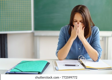 Tired Teacher Or Mature Female Student Sitting At A Desk In The Classroom With Paperwork Holding Her Hands To Her Face