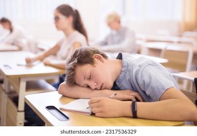 Tired teen student sleeping at desk in classroom during lesson with blurred classmates in background - Powered by Shutterstock