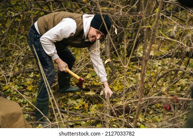 Tired tourist male wearing warm clothes chopping firewood with axe in forest on overcast cold day. Concept of survival, travel, hiking, camping in nature.