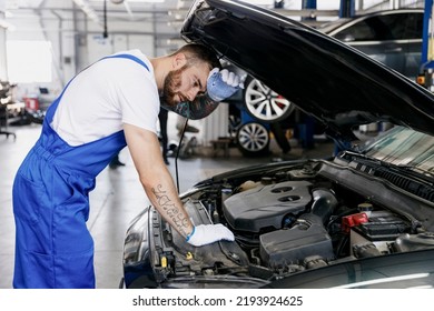 Tired Troubleshooter Young Male Professional Technician Car Mechanic Man In Denim Blue Overalls White T-shirt Fixing Problem With Raised Hood Work In Light Modern Vehicle Repair Shop Workshop Indoor