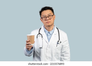 Tired Unhappy Middle Aged Chinese Male Doctor In White Coat And Glasses With Cup Of Coffee Takeaway, Isolated On Blue Background. Fatigue, Overwork, Problems With Medicine, Treatment And Health Care