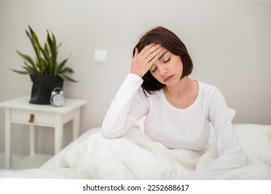 Tired unhappy young beautiful brunette woman waking up with headache in the morning, sick lady sitting in bed at home, touching her head, suffering from hangover or migraine, copy space