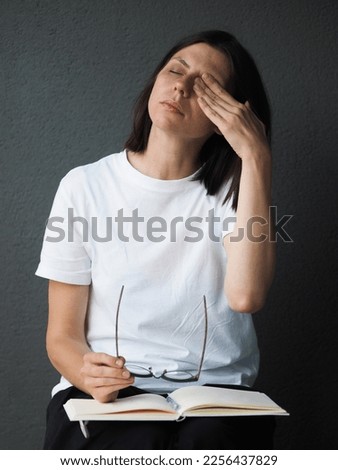 Tired upset brunette woman, studet or manager or freelancer, in white t-shirt, tired of work, experiencing stress, taking break from work, rubs her eyes with her hand, has headache. Unhappy, worried.
