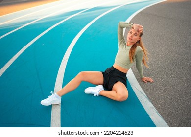 Tired Upset Female Athlete Sitting On Running Track Resting After Jogging Exercises. Disappointed Sport Result. Amateur Sprinter Sportswoman Exhausted After Tough Trainings Sitting On Track