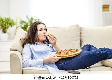 Tired woman is chewing pizza, while laying on the white sofa. She is watching TV shows, being on blurred background.
