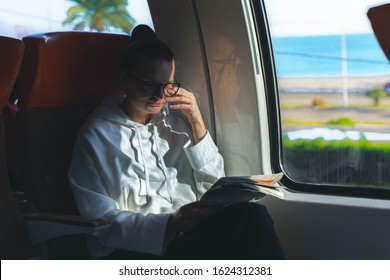 Tired Woman In Eyeglasses Rubbing Eyes And Sitting In Train Near Big Window And Holding Newspaper. Woman With Closed Eyes. Sea And Palms Outside Train Window.