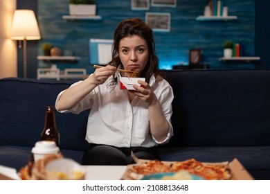 Tired woman holding chopsticks and chinese takeaway noodle box sitting on couch after long day at the office. Person looking sad eating delivery asian fast food watching evening news.