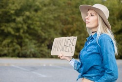 Tired Woman Hopefully Look Out Passing Cars With Cardboard Poster On Roadside In Forest. Lady In Hat And Denim Outfit Escape From City To Go Anywhere. Travelling, Freedom, Hitchhiking, Vacations.