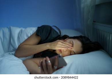 Tired woman lying in bed sleepless holding smartphone in hand reading about sleep disorder and mental health in dark bedroom. Anxious female need rest. Sleeplessness, anxiety and depression concept