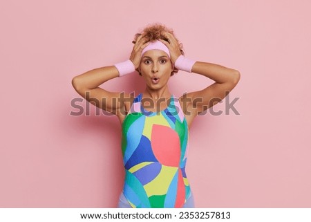 Tired woman posing on pink backdrop, wearing multycoloured leotard, opened mouth, hands pressed to her head, lifestyle concept, copy space.