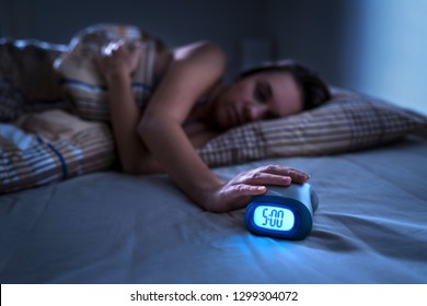 Tired woman waking up for work or school early in the morning. Grumpy lady pushing snooze button or turning off alarm clock with hand. Getting up after sleeping. Exhausted person in bed. Dark bedroom. - Shutterstock ID 1299304072