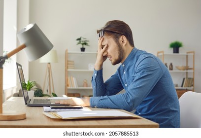Tired worker having problem. Man makes dumb mistake or has difficulty with digital file. Side view of upset stressed young guy facepalming sitting at working desk with laptop computer in home office