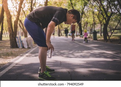 a tired young Asian male resting holding a bottle of drinking water in the road at the park after a daily exercise, running jogging for his fitness in the warm summer morning