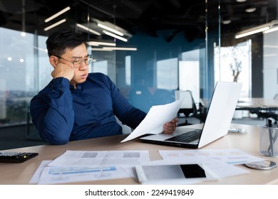 Tired young Asian man, accountant, businessman, freelancer. He sits in the office at a table with a laptop, looks through documents, holds his head in his hands, sighs a hard and boring day. - Powered by Shutterstock