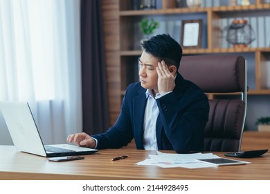 Tired young asian man businessman, worker, lawyer at work. He has a headache, keeps his hands on his head, sits at a desk in the office