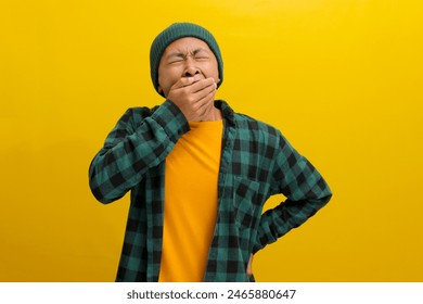 Tired young Asian man, wearing a beanie hat and casual outfit, yawns wearily, likely due to insomnia or being overworked. He covers his mouth with his hand while standing against a yellow background - Powered by Shutterstock