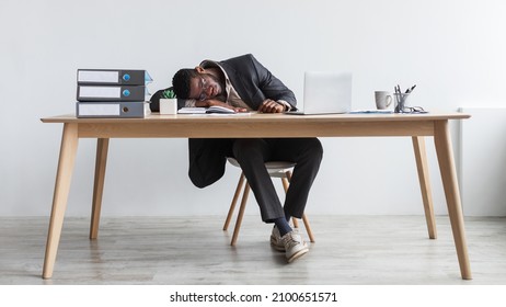 Tired young black businessman sleeping on his desk in front of laptop, overworking, feeling exhausted at office, full length. Millennial African American employee suffering from fatigue at workplace