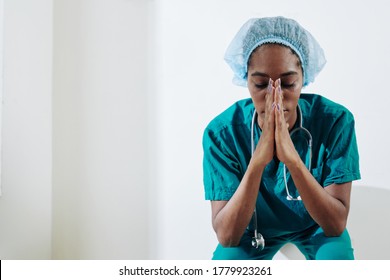 Tired young Black medical nurse praying for sick and suffering people when sitting on the floor in hospital corridor - Powered by Shutterstock