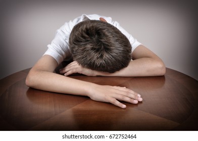 Tired young boy sitting and sleeping with his head and hands on the brown table 