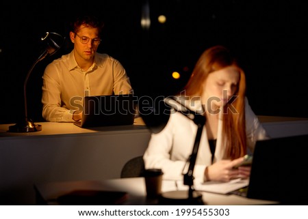 Tired Young Capable Manager Working Late Hours in Office. Office is Dark. Male is working on computer with colleagues. business, deadline, work concept. focus on man