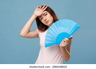 Tired Young Caucasian Lady With Fan Touching Her Sweaty Forehead, Feeling Exhausted Because Of Summer Heat, Suffering From High Temperature Outside, Blue Studio Background, Copy Space.