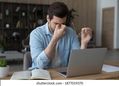 Tired young Caucasian man work on laptop at home office take off glasses overwhelmed with computer job. Exhausted male employee suffer from headache or dizziness from screen. Eyesight problem concept. - Shutterstock ID 1877019370
