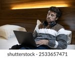 Tired young entrepreneur man on business trip works in the hotel room with his laptop on the bed, listening to music with headphones while working
