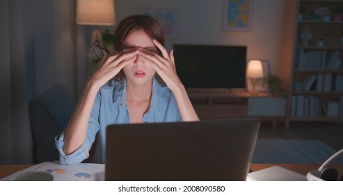 Tired Young Freelance Worker Rubbing Her Eyes While Sitting In Front Of Laptop Computer And Working On Promising Project - Asian Girl Soho Lady Stay Up Late In Home Office Holding Glasses Eye Painful