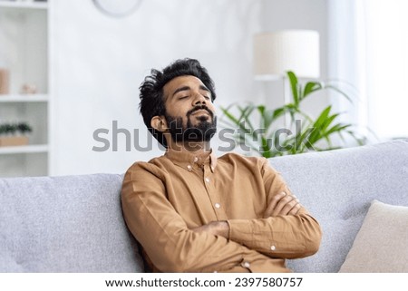 Tired young Indian man napping on couch at home with arms folded on chest, hard day's work, rest.