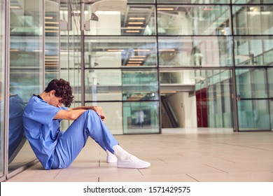 Tired Young Male Doctor Wearing Scrubs Sitting Against Wall In Modern Hospital Building - Powered by Shutterstock