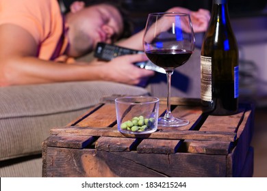 Tired Young Man, Bachelor Sleeping On A Couch  During Watching Movie, Series, Tv Show In The Late Evening, Night. Wine And Unhealthy Snacks On A Table.