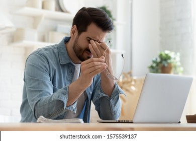 Tired young man feel pain eyestrain holding glasses rubbing dry irritated eyes fatigued from computer work, stressed man suffer from headache bad vision sight problem sit at home table using laptop