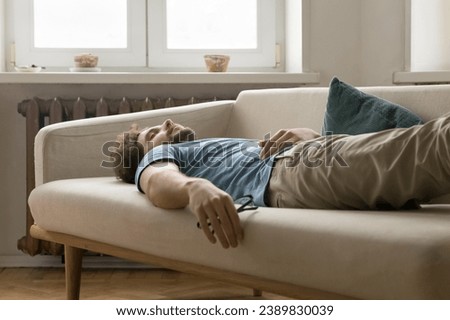 Tired young man having daytime nap on sofa at home. Overloaded guy in casual wear falling asleep on couch in living room, take break, relieving exhaustion after hard working day. Lack of energy, rest