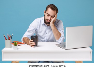 Tired Young Man In Light Shirt Sit Work At Desk With Pc Laptop Isolated On Blue Background. Achievement Business Career Concept. Mock Up Copy Space. Put Hand On Cheek Sleep Hold Cup Of Coffee Or Tea