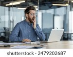 Tired young man, office worker sits in the office overtime, works on a laptop, yawns, covers his mouth with his hand.