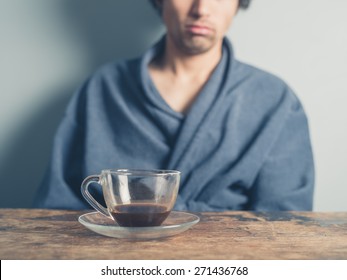 A Tired Young Man Wearing A Bathrobe Is Sitting At A Table And Is Trying To Wake Up By Having A Cup Of Coffee