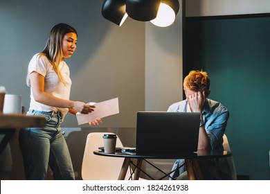 Tired Young Man Working On A Computer Bowed His Head, Not Paying Attention To A Colleague With Papers.