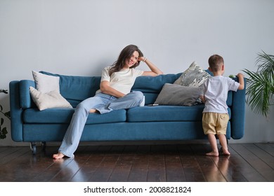 Tired Young Mother Or Nanny Sits On The Couch And Watches Her Son Playing.